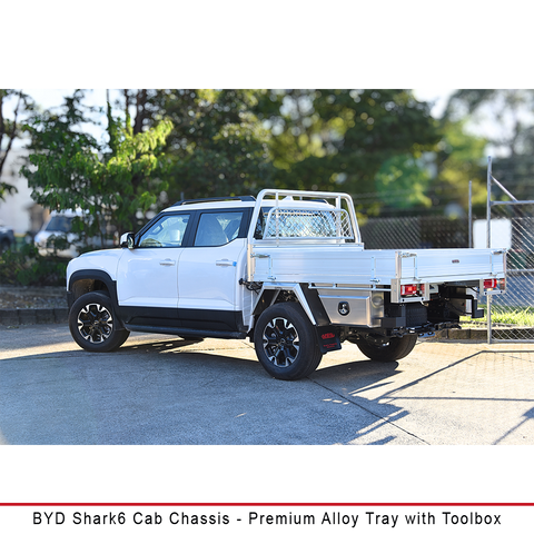 A white BYD Shark6 cab chassis truck with a premium silver alloy tray and toolbox, parked outdoors on a paved surface with trees and a fence in the background.