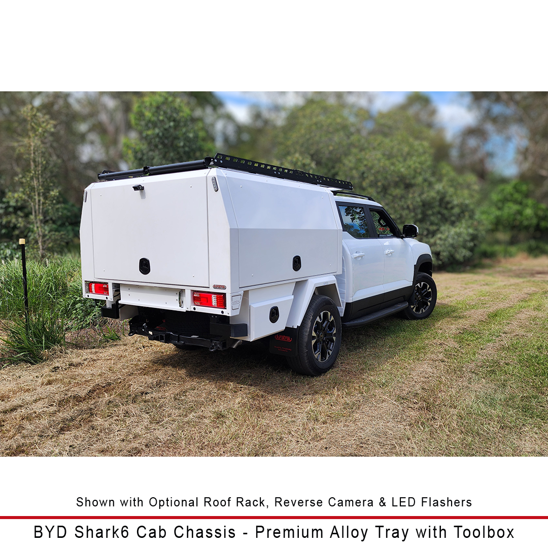 A white BYD Shark6 cab chassis truck fitted with a premium alloy roof tray and toolbox, parked outdoors on a grassy path with trees in the background. Text at the bottom describes the optional roof rack, reverse camera, LED flashers, and identifies the vehicle model.