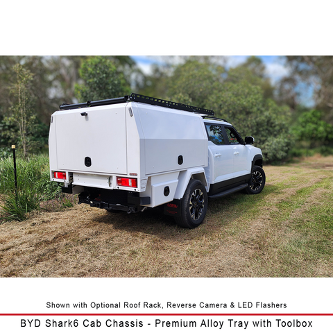 A white BYD Shark6 cab chassis truck fitted with a premium alloy roof tray and toolbox, parked outdoors on a grassy path with trees in the background. Text at the bottom describes the optional roof rack, reverse camera, LED flashers, and identifies the vehicle model.